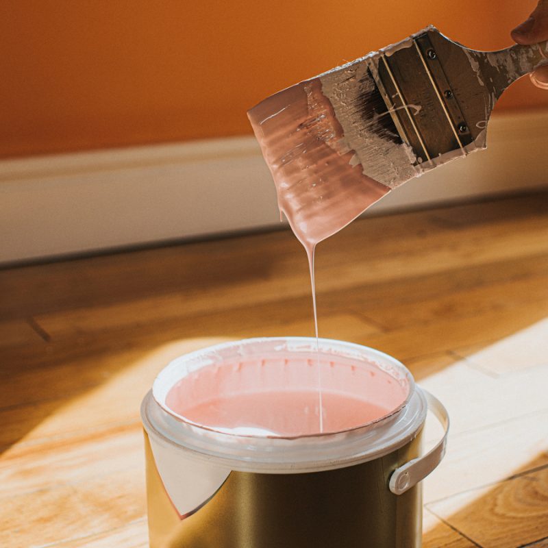 Hand holds a thick paint brush and removes it, after dipping it in the paint. It creates a droplet of paint which splashes back into the pot. The wall behind is painted orange and the colour in the tin is pink, which clashes. Conceptual with space for copy.
